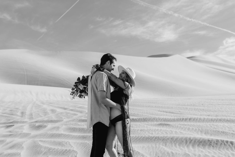 black and white photo of couple holding and looking at each other while holding bridal bouquet during maternity session in nevada sand dunes by katherine krakowski photography a lake tahoe reno photographer