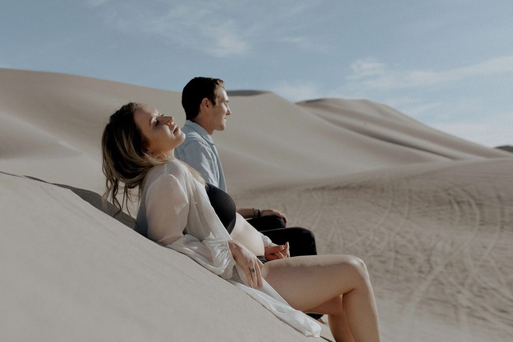 couples laying on sand in desert in sand dunes in nevada during maternity session in lingerie by katherine krakowski photography a lake tahoe reno photographer