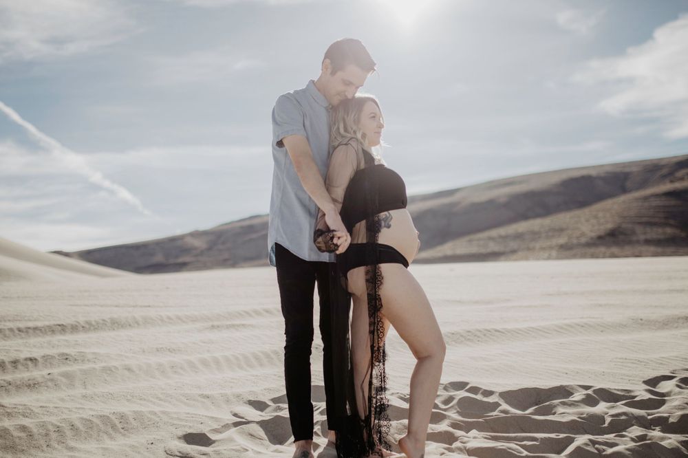 couple holding hands and kissing head wearing black lingerie in desert sand dunes during maternity session by katherine krakowski photography a lake tahoe reno photographer