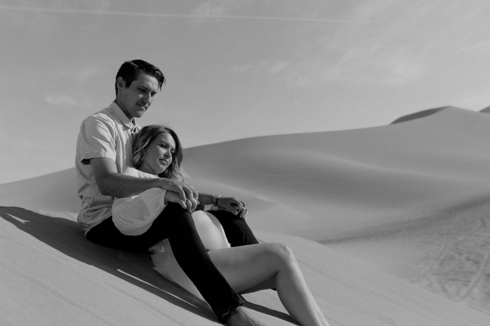 couples laying on sand in desert in sand dunes in nevada during maternity session in lingerie by katherine krakowski photography a lake tahoe reno photographer