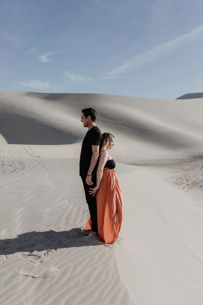 couple standing back to back wearing black and orange maxi skirt in desert sand dunes during maternity session by katherine krakowski photography a lake tahoe reno photographer