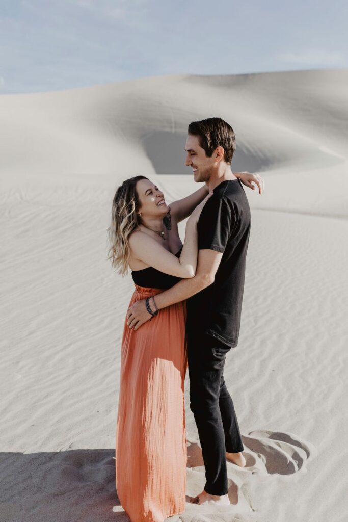 couple holding each other and laughing wearing black and orange maxi skirt in desert sand dunes during maternity session by katherine krakowski photography a lake tahoe reno photographer