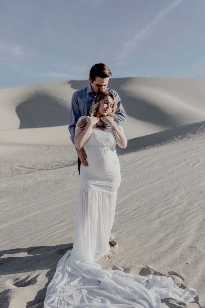 couple in wedding gown attire holding each other  in desert sand dunes during maternity session by katherine krakowski photography a lake tahoe reno photographer