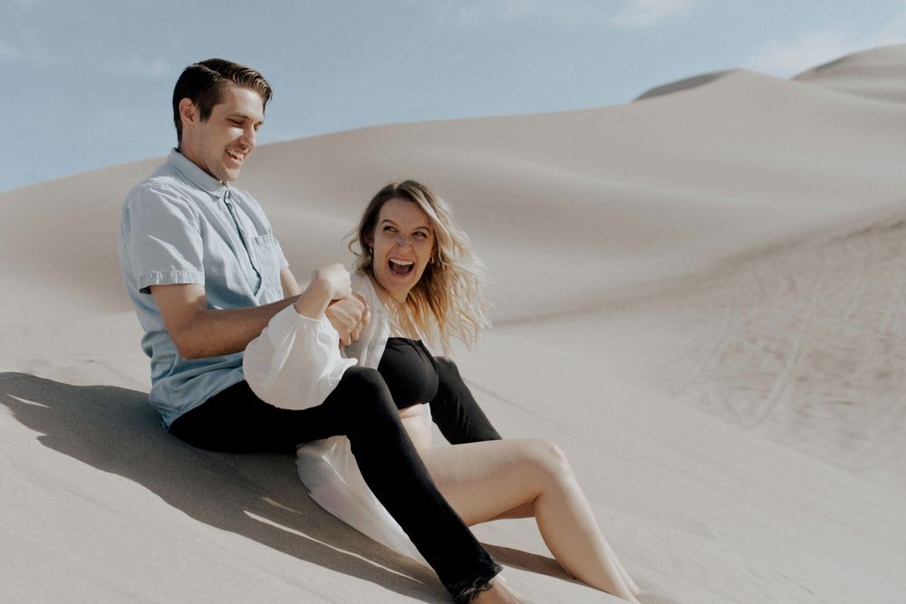 couples sitting and laughing on sand in desert in sand dunes in nevada during maternity session in lingerie by katherine krakowski photography a lake tahoe reno photographer