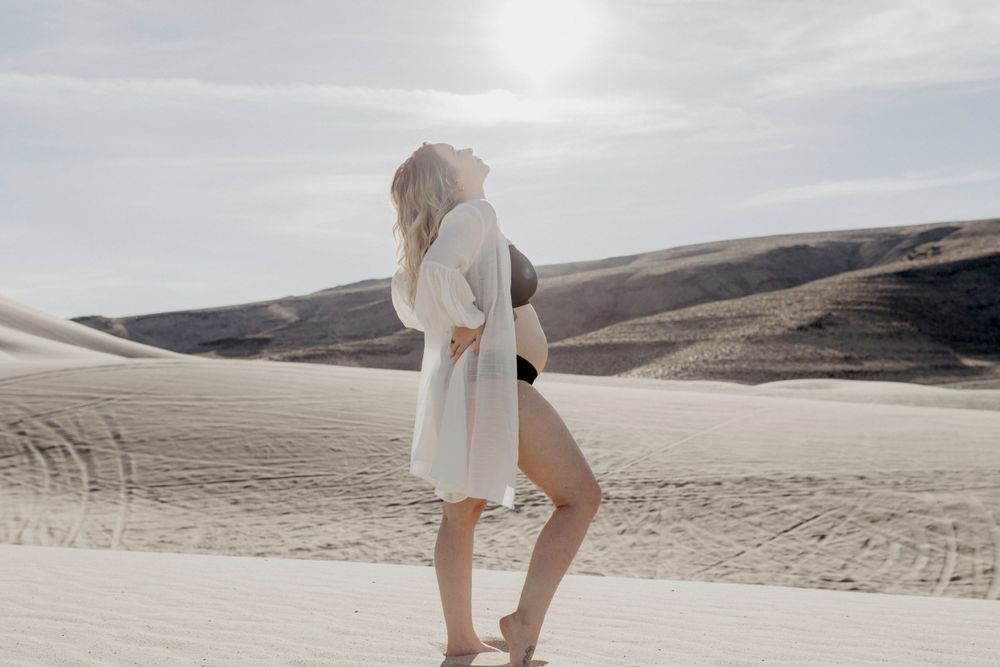 pregnant female in black lingerie and white oversized shirt standing in front of rising sun in the nevada desert sand dunes by katherine krakowski photography a lake tahoe reno photographer