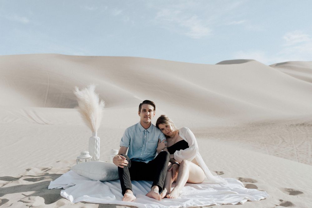 couples sitting on blanket holding each other with pampass grass decor sand in desert in sand dunes in nevada during maternity session in lingerie by katherine krakowski photography a lake tahoe reno photographer