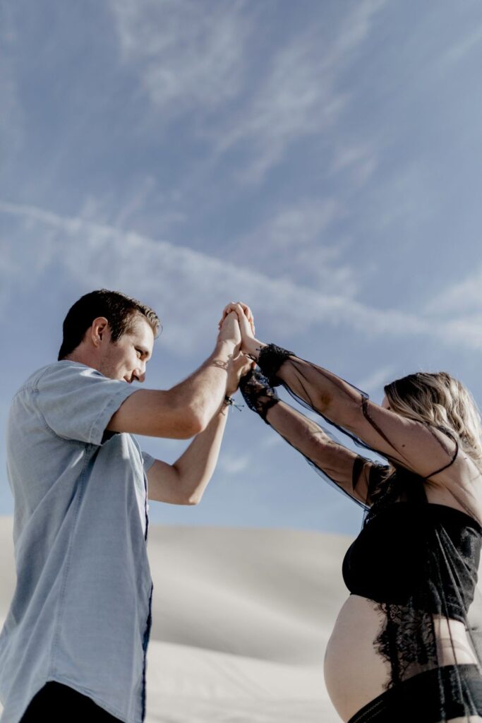underneath photo of couple holding hands in sky wearing black lingerie in desert sand dunes during maternity session by katherine krakowski photography a lake tahoe reno photographer