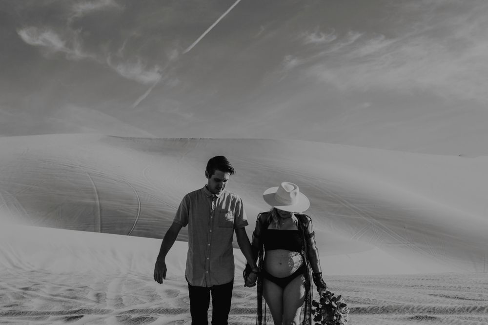 couple holding hands looking down holding bouquet during maternity session wearing black lingerie and wide brim hat in nevada sand dunes by katherine krakowski photography a lake tahoe reno photographer