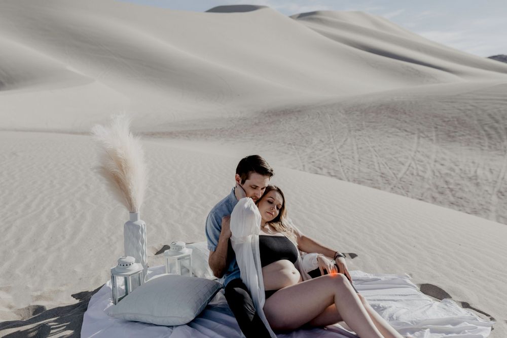 couples sitting on blanket holding each other with pampass grass decor sand in desert in sand dunes in nevada during maternity session in lingerie by katherine krakowski photography a lake tahoe reno photographer