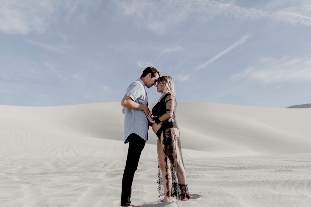 couple holding hands with foreheads together wearing black lingerie in desert sand dunes during maternity session by katherine krakowski photography a lake tahoe reno photographer