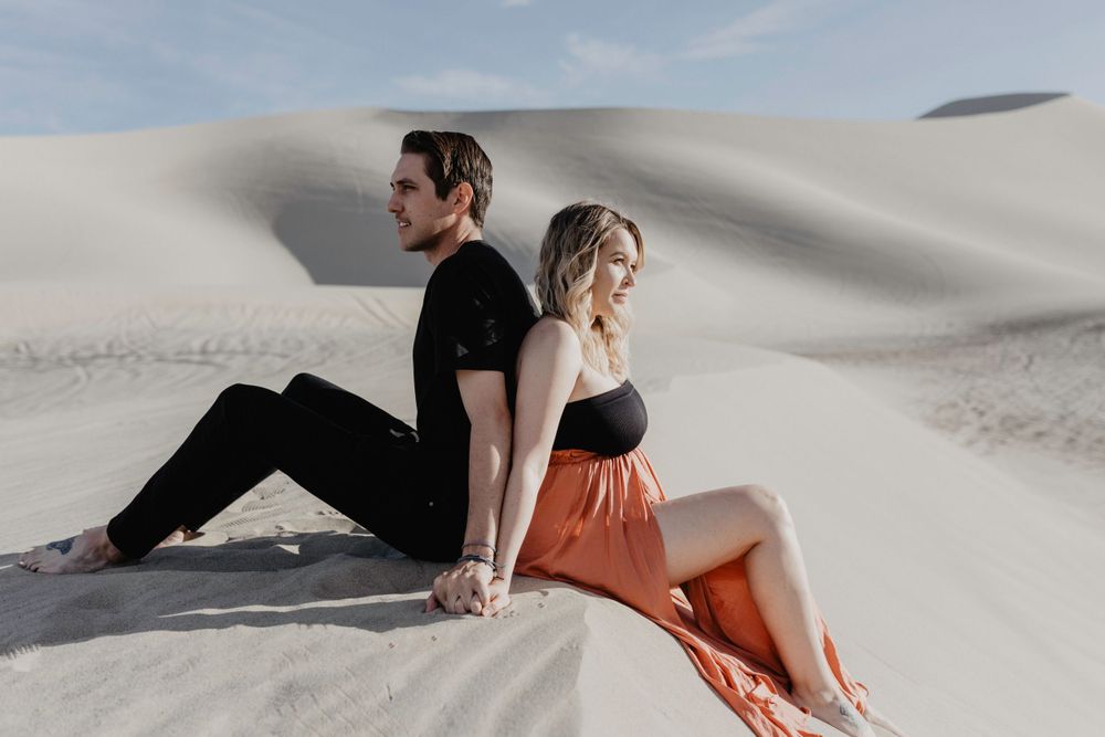 couple standing back to back wearing black and orange maxi skirt in desert sand dunes during maternity session by katherine krakowski photography a lake tahoe reno photographer