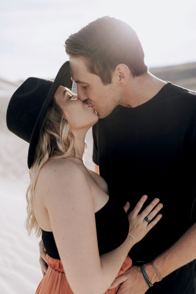 couples close up kissing each other wearing black and wide brim hat in sand dunes in nevada 