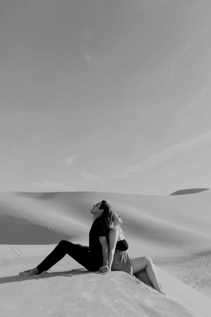 couple sitting back to back wearing black lingerie in desert sand dunes during maternity session by katherine krakowski photography a lake tahoe reno photographer