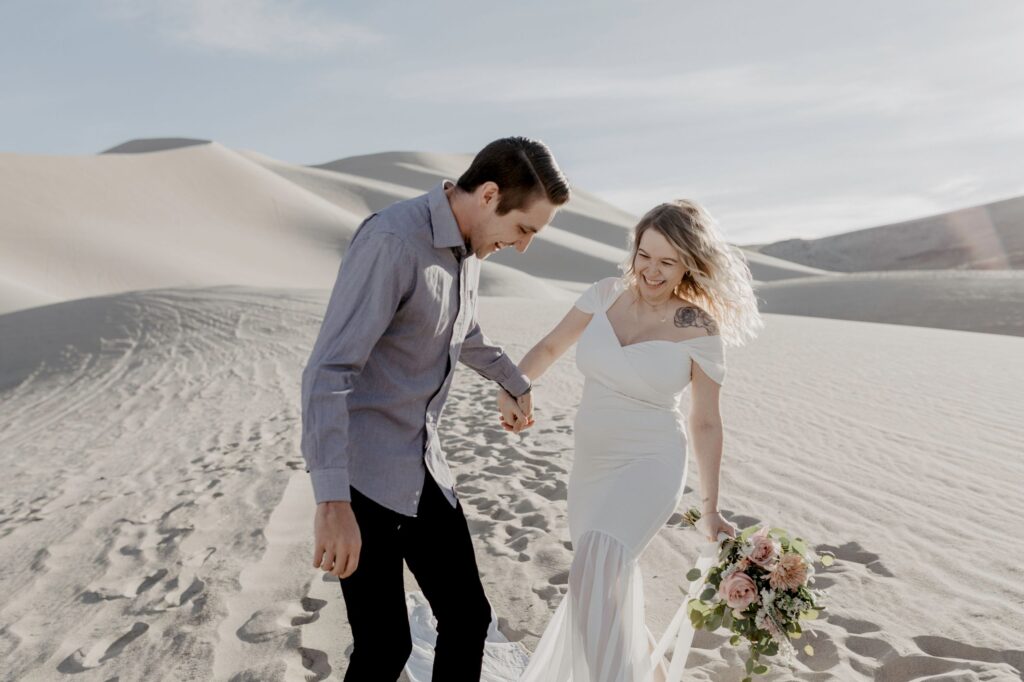 couple holding each other and laughing wearing wedding dress in desert sand dunes during maternity bridal session by katherine krakowski photography a lake tahoe reno photographer