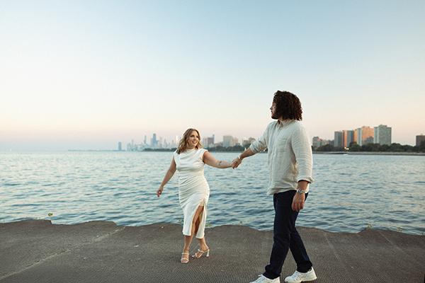 Jackie and Randy walking hand in hand along Lake Michigan during their Montrose Harbor engagement session in Chicago with the skyline in the background